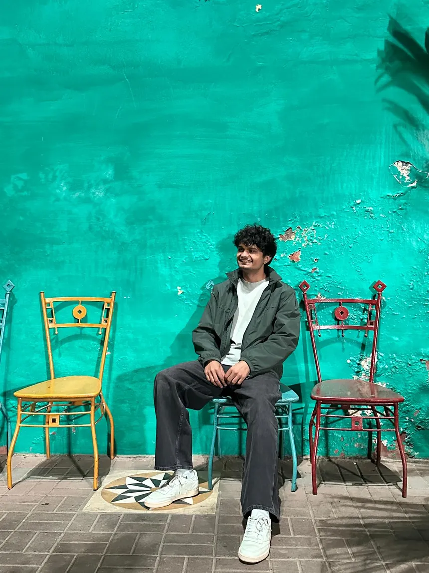 Issa sitting against a bright green wall between mismatched colorful chairs